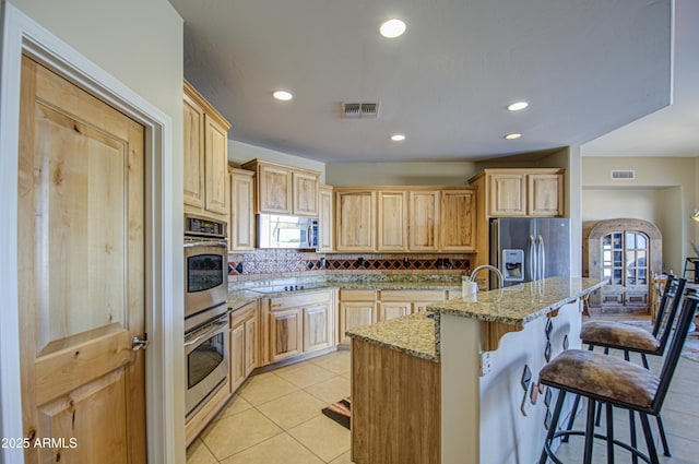 kitchen with a breakfast bar, light brown cabinetry, stainless steel appliances, light stone countertops, and a center island with sink