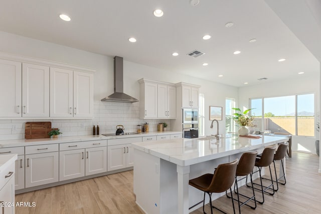 kitchen featuring a center island with sink, oven, wall chimney exhaust hood, black electric cooktop, and white cabinetry