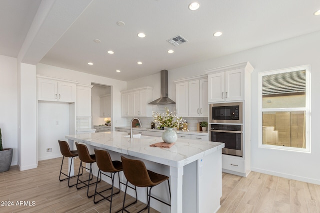 kitchen with a center island with sink, oven, white cabinetry, and wall chimney range hood