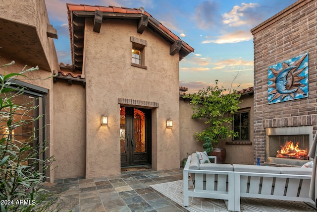 exterior entry at dusk with an outdoor brick fireplace, stucco siding, a patio area, and a tiled roof