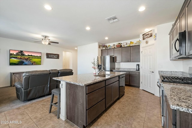 kitchen featuring black appliances, an island with sink, sink, dark brown cabinetry, and light stone counters