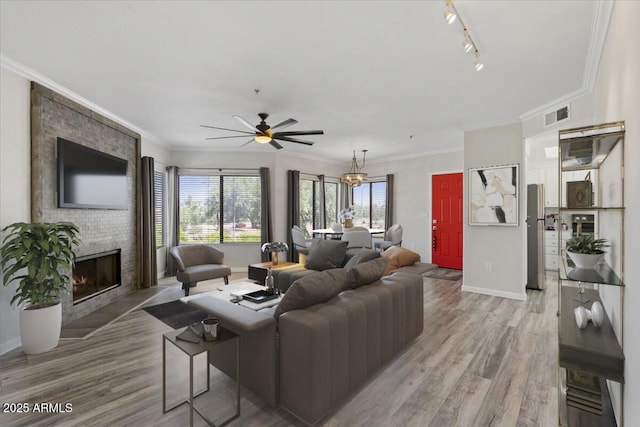 living area with ornamental molding, ceiling fan with notable chandelier, light wood finished floors, a brick fireplace, and baseboards