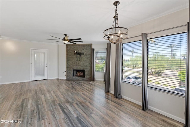 unfurnished living room with a fireplace, crown molding, baseboards, wood finished floors, and ceiling fan with notable chandelier
