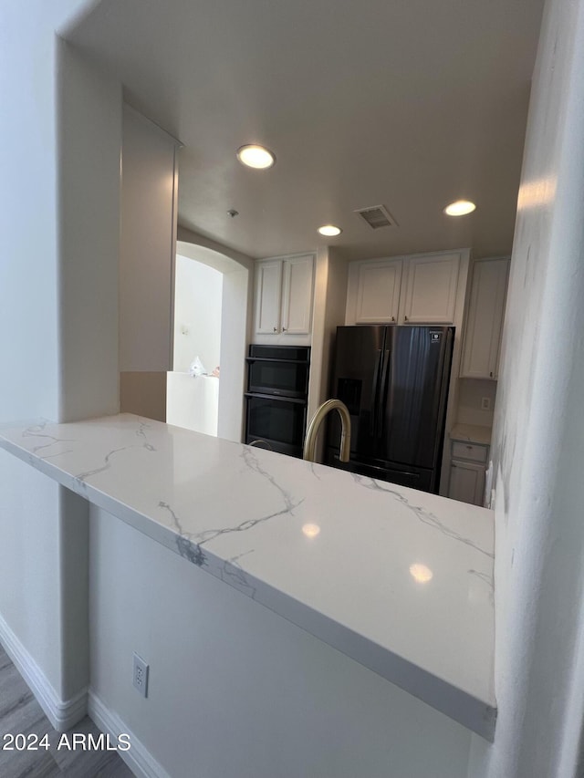 kitchen with white cabinetry, stainless steel fridge with ice dispenser, kitchen peninsula, black double oven, and light stone countertops