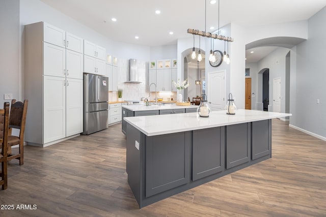 kitchen featuring wall chimney exhaust hood, a spacious island, pendant lighting, white cabinets, and stainless steel refrigerator