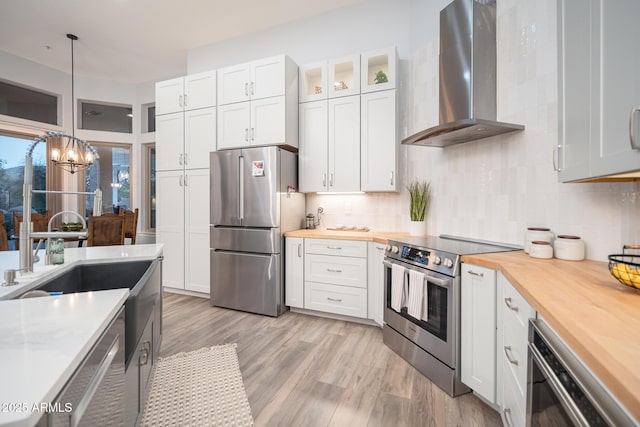 kitchen featuring wooden counters, appliances with stainless steel finishes, wall chimney range hood, white cabinetry, and hanging light fixtures