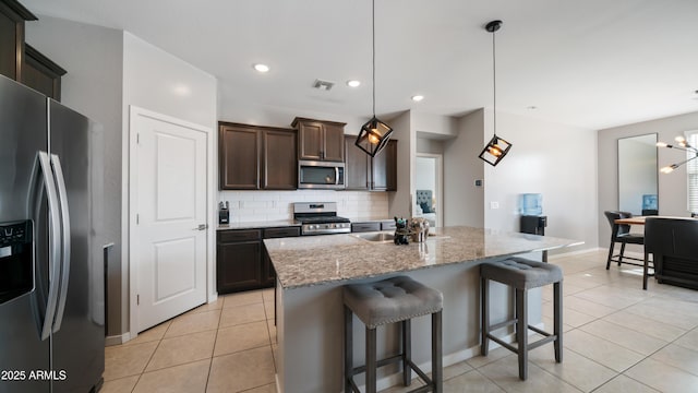 kitchen with light stone counters, dark brown cabinets, hanging light fixtures, stainless steel appliances, and a kitchen island with sink