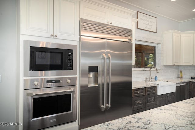 kitchen featuring backsplash, white cabinetry, a sink, light stone countertops, and built in appliances