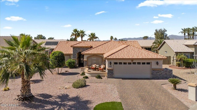 mediterranean / spanish house with an attached garage, stucco siding, stone siding, a tiled roof, and decorative driveway