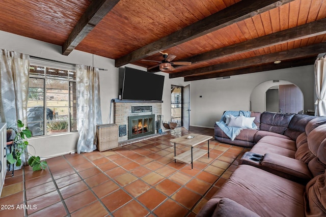 tiled living room featuring beam ceiling, a ceiling fan, wooden ceiling, baseboards, and a brick fireplace