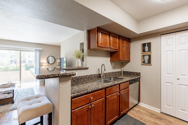 kitchen featuring dark stone countertops, a sink, light wood-style floors, stainless steel dishwasher, and brown cabinets