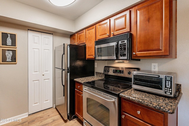 kitchen with brown cabinetry, dark stone counters, a toaster, appliances with stainless steel finishes, and light wood-type flooring