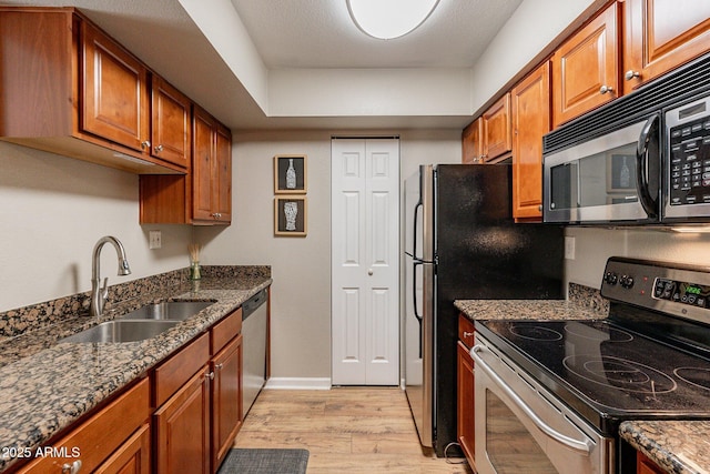 kitchen with light wood finished floors, dark stone counters, brown cabinets, stainless steel appliances, and a sink