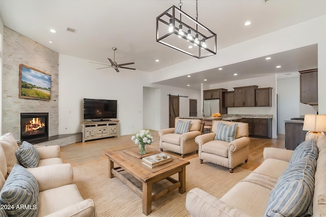 living room featuring light wood-type flooring, ceiling fan with notable chandelier, a barn door, and a stone fireplace