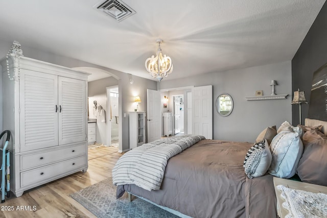 bedroom with light wood finished floors, visible vents, arched walkways, ensuite bathroom, and a chandelier