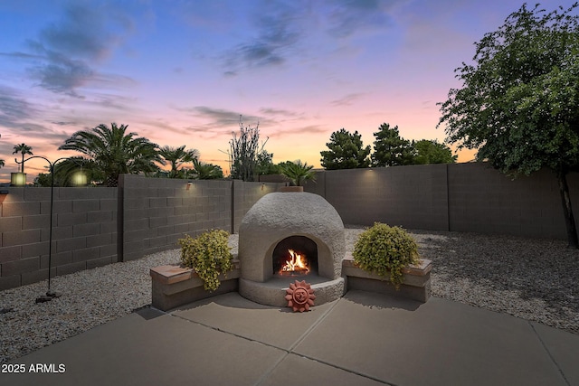 patio terrace at dusk featuring a warm lit fireplace and a fenced backyard