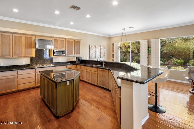 kitchen with pendant lighting, hardwood / wood-style flooring, appliances with stainless steel finishes, a kitchen island, and wall chimney exhaust hood