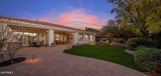 back house at dusk featuring a patio and a lawn