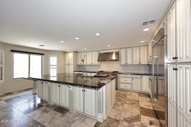 kitchen featuring exhaust hood, stone tile flooring, recessed lighting, cream cabinetry, and baseboards