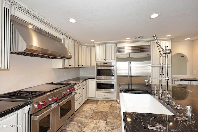 kitchen with high quality appliances, ventilation hood, cream cabinetry, recessed lighting, and light stone finish flooring