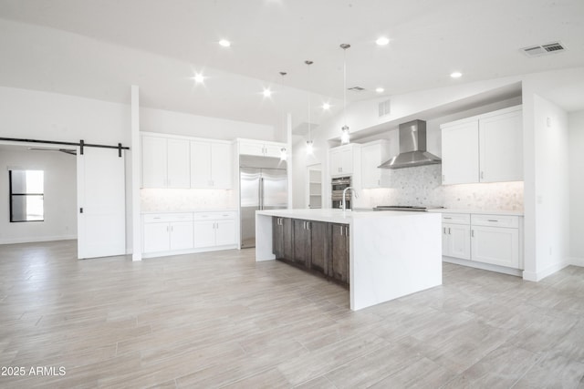 kitchen featuring a center island with sink, vaulted ceiling, wall chimney exhaust hood, a barn door, and decorative light fixtures