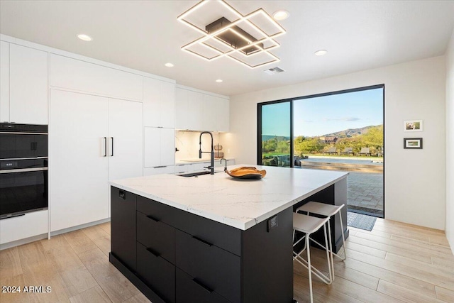 kitchen featuring sink, an island with sink, double oven, white cabinetry, and light wood-type flooring