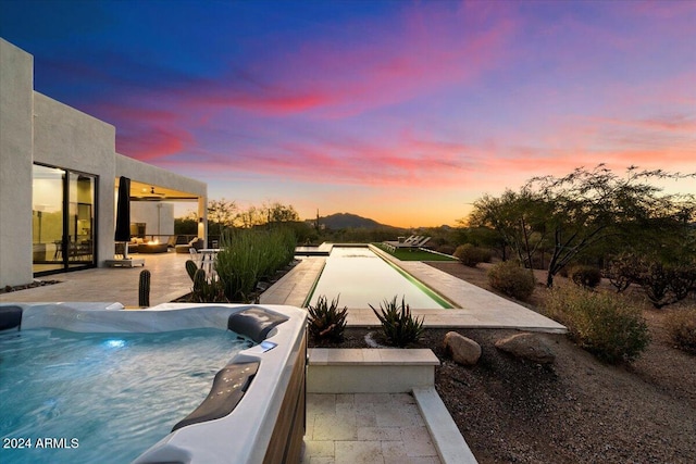 pool at dusk with a hot tub, a mountain view, and a patio