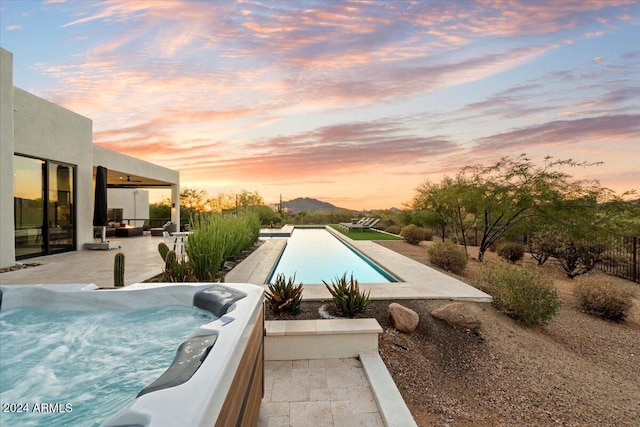 pool at dusk with ceiling fan, a mountain view, a patio, and a hot tub