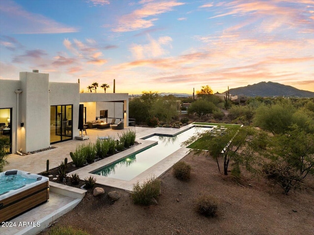 pool at dusk with a patio, outdoor lounge area, a mountain view, and an outdoor hot tub