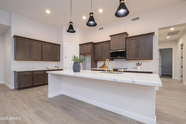 kitchen with light stone counters, dark brown cabinetry, sink, decorative light fixtures, and a center island with sink