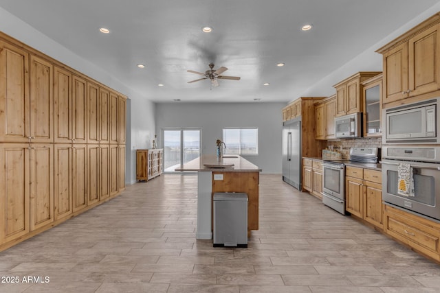 kitchen featuring an island with sink, built in appliances, ceiling fan, sink, and backsplash