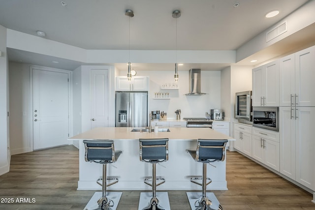 kitchen with appliances with stainless steel finishes, decorative light fixtures, white cabinetry, wall chimney range hood, and a center island with sink