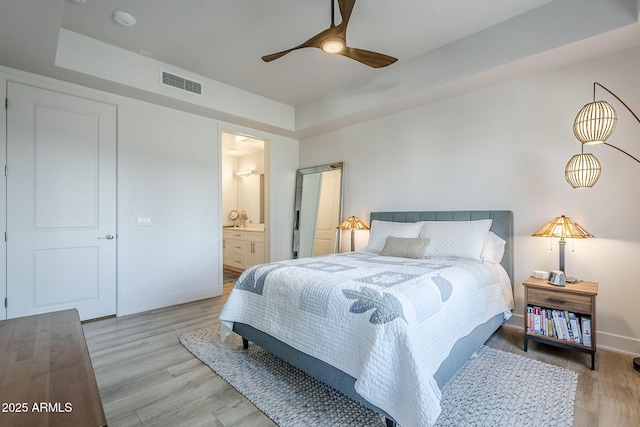 bedroom with ensuite bath, a raised ceiling, and light wood-type flooring