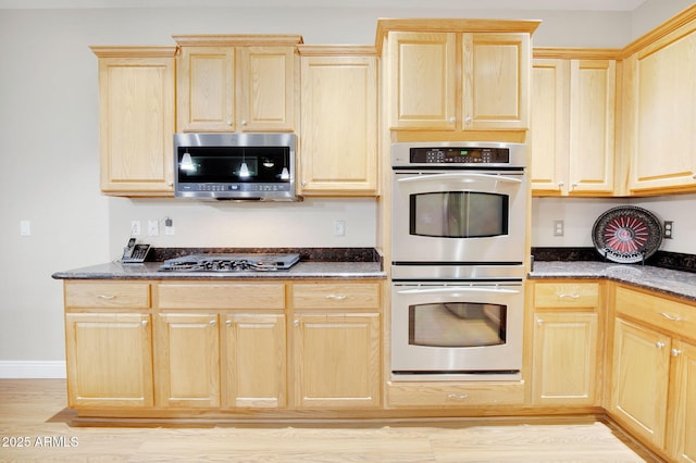 kitchen with baseboards, light wood-style floors, appliances with stainless steel finishes, light brown cabinetry, and dark stone countertops