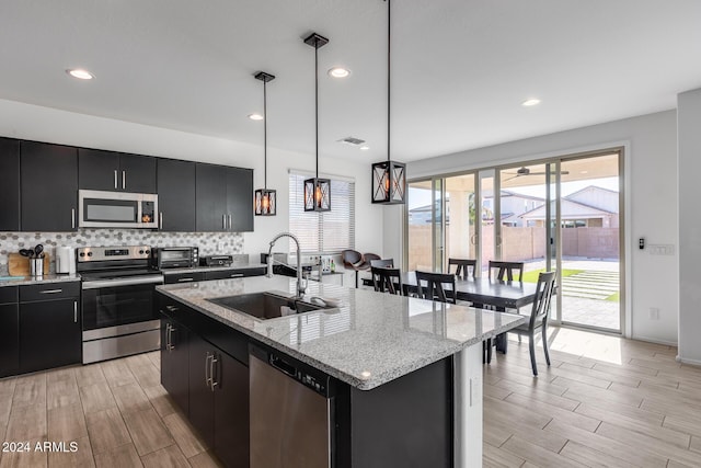 kitchen with appliances with stainless steel finishes, a kitchen island with sink, sink, decorative light fixtures, and light hardwood / wood-style floors