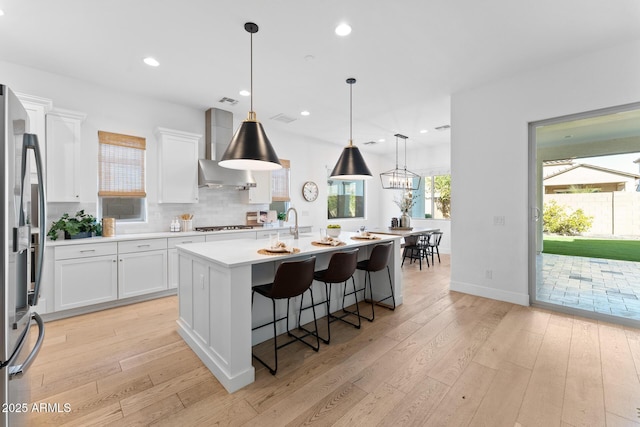 kitchen featuring white cabinetry, wall chimney exhaust hood, decorative light fixtures, and a center island with sink