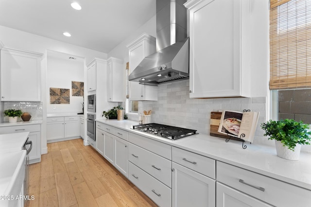 kitchen featuring wall chimney range hood, light hardwood / wood-style flooring, white cabinetry, stainless steel appliances, and tasteful backsplash