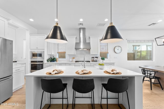 kitchen with hanging light fixtures, appliances with stainless steel finishes, wall chimney range hood, a kitchen island with sink, and white cabinets