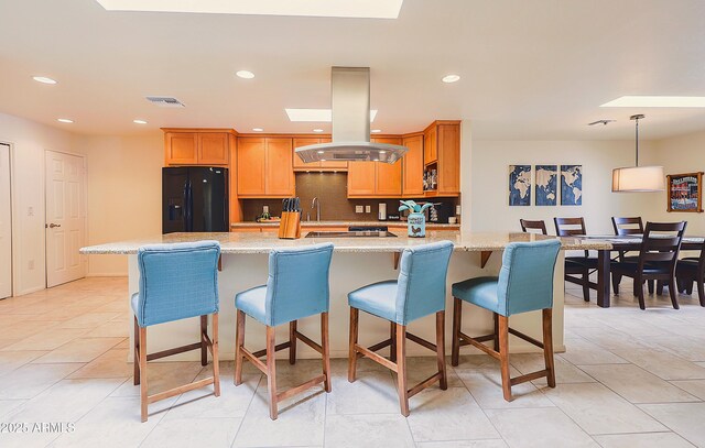 kitchen with black refrigerator with ice dispenser, visible vents, island exhaust hood, a center island with sink, and a skylight
