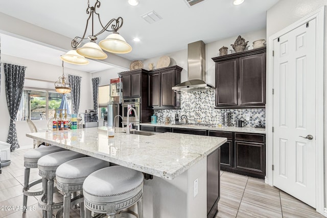 kitchen with decorative light fixtures, wall chimney range hood, stainless steel appliances, a kitchen island with sink, and a breakfast bar area