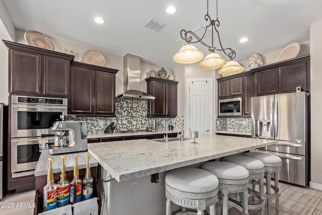 kitchen featuring appliances with stainless steel finishes, sink, backsplash, a kitchen island with sink, and wall chimney range hood