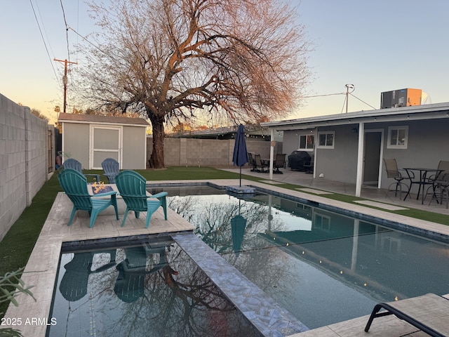 pool at dusk with an outbuilding, a patio area, a fenced backyard, and a shed