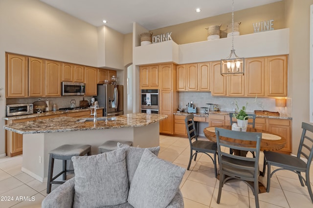 kitchen with a notable chandelier, a kitchen island with sink, hanging light fixtures, stainless steel appliances, and light stone counters