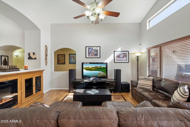 living area featuring baseboards, ceiling fan, wood finished floors, a glass covered fireplace, and high vaulted ceiling