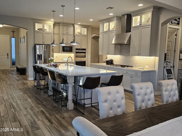 kitchen featuring stainless steel appliances, arched walkways, wall chimney range hood, and a kitchen breakfast bar