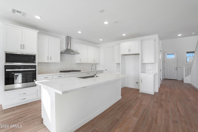 kitchen with appliances with stainless steel finishes, light wood-type flooring, sink, wall chimney range hood, and a center island with sink