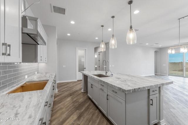 kitchen featuring decorative backsplash, a kitchen island with sink, pendant lighting, wall chimney exhaust hood, and sink