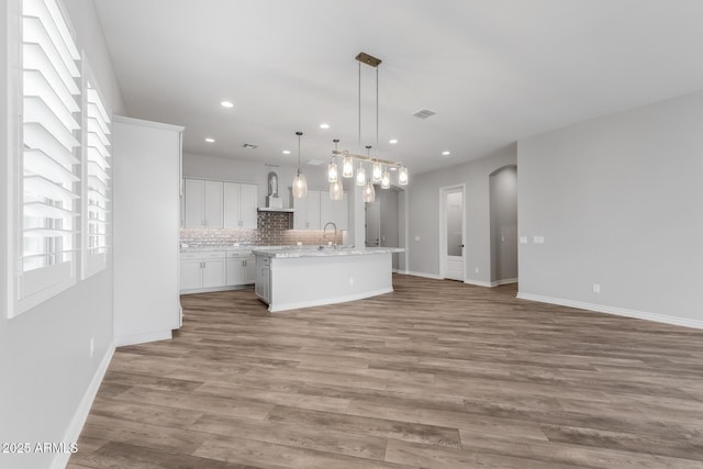 kitchen featuring white cabinets, wall chimney range hood, an island with sink, decorative backsplash, and hanging light fixtures