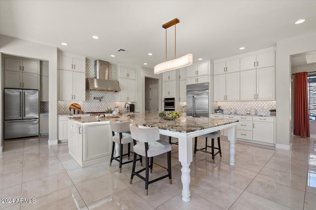 kitchen featuring a spacious island, wall chimney exhaust hood, light stone countertops, appliances with stainless steel finishes, and a breakfast bar area