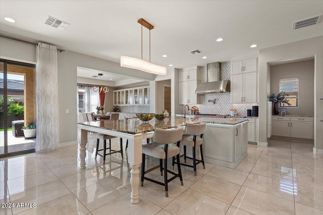 kitchen with pendant lighting, wall chimney exhaust hood, light stone countertops, and an island with sink